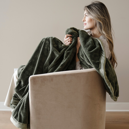 A woman sitting in a chair is holding a green lush blanket. This luxury blanket is a soft blanket and a Saranoni blanket. This extra large throw blanket is great for gifting, relaxing, or as a home decor staple. Enjoy this cozy blanket for years to come. 