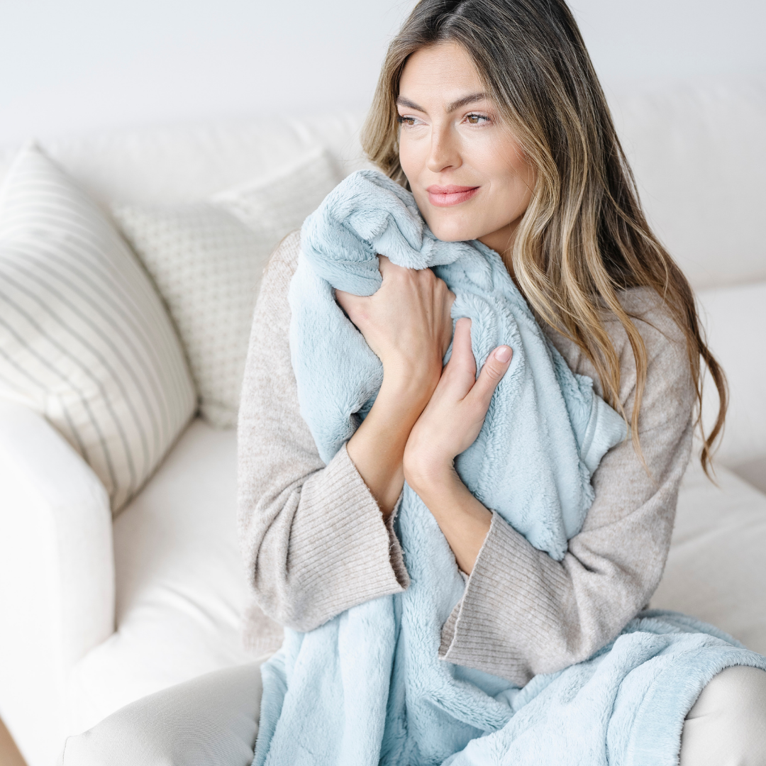 A woman sitting on a couch is hugging a light blue blanket that is a lush blanket. This Saranoni blanket is so soft and is also a luxury blanket. This extra large blanket is great for gifting, relaxing, or adding to your home decor. 