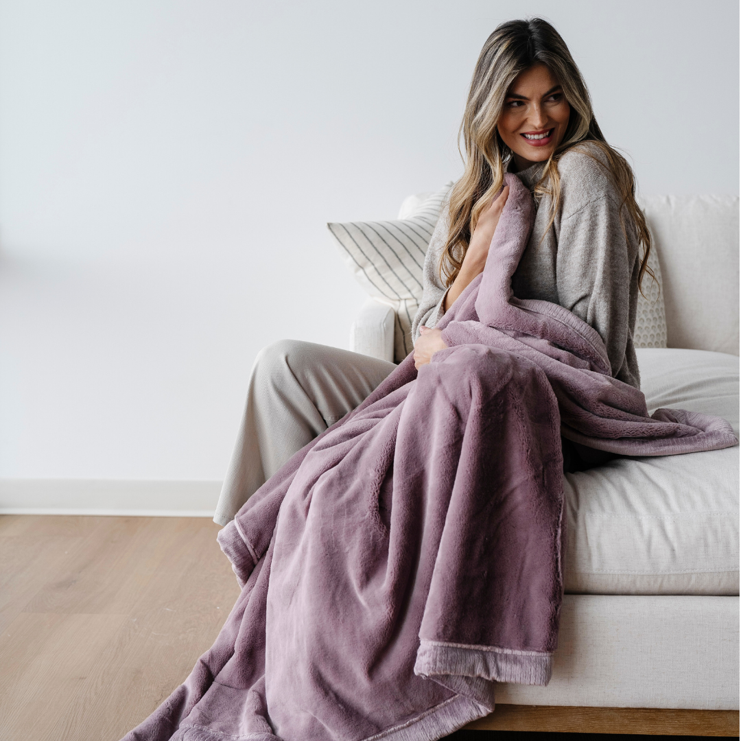 A smiling woman is sitting on a couch and holding a light purple lush blanket. This soft blanket is a luxury blanket and a Saranoni blanket. Use this extra large blanket as a comfort gift, home decor, or a relaxation blanket. 