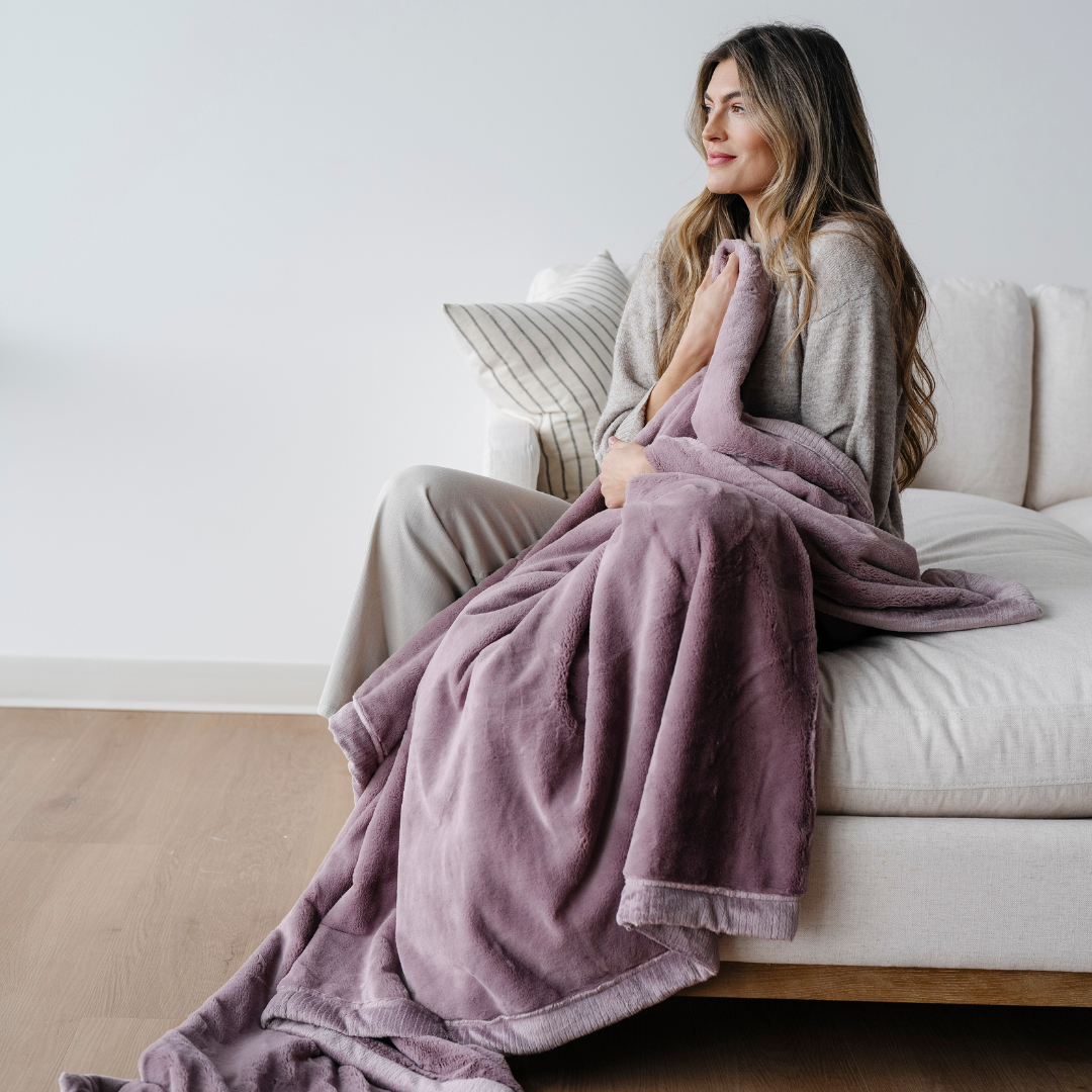 A woman sitting on a couch holds a light purple blanket. This luxury blanket is a soft blanket and a Saranoni blanket. This extra large blanket is a great gift or for relaxing. Enjoy this coziness for years to come. 