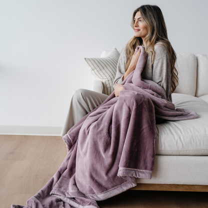 A woman sitting on a couch holds a light purple blanket. This luxury blanket is a soft blanket and a Saranoni blanket. This extra large blanket is a great gift or for relaxing. Enjoy this coziness for years to come. 