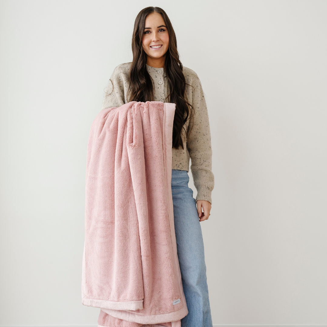 A woman smiles and holds an extra large blanket that is a pink lush blanket. This luxury blanket is a soft blanket and a Saranoni blanket. Use this blanket for gifting, relaxing, or as a home decor staple. Enjoy this cozy blanket for years to come!