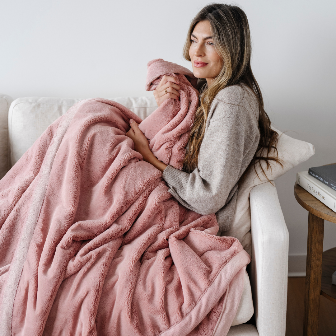A woman is sitting on a couch. She is holding a lush blanket that is a pink blanket. This extra large blanket is a luxury blanket and a Saranoni blanket. This blanket is a great gift for her and is also great for home decor. Enjoy this faux fur blanket for years to come.
