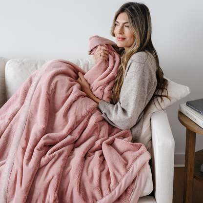A woman is sitting on a couch. She is holding a lush blanket that is a pink blanket. This extra large blanket is a luxury blanket and a Saranoni blanket. This blanket is a great gift for her and is also great for home decor. Enjoy this faux fur blanket for years to come.
