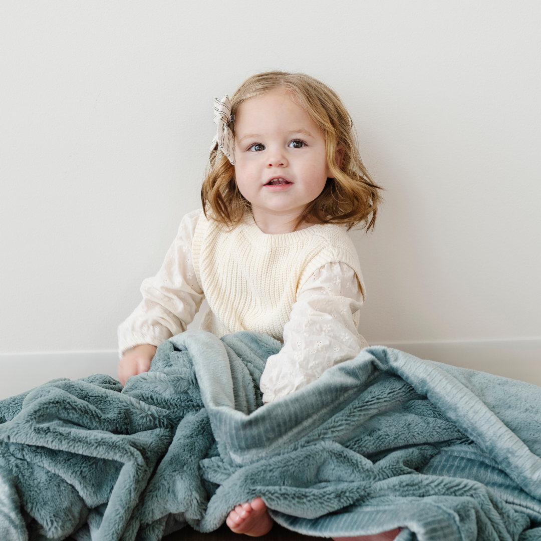 A little girl is sitting with a eucalyptus lush blanket. This soft blanket is lush blanket and a Saranoni blanket. A cozy addition to a nursery or bedroom, this receiving blanket is made to last and provide the ultimate comfort for your little one. 