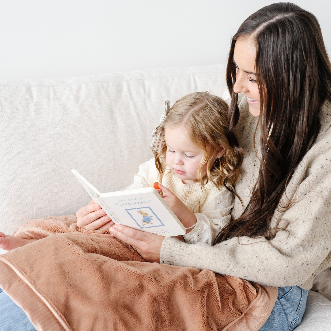 A mother reads to her little girl while they are sitting on a couch. On their laps is a buttery-soft Saranoni blanket that is a luxury blanket. This is a great baby gift of gift for kids and is durable and cozy. Enjoy this soft blanket!
