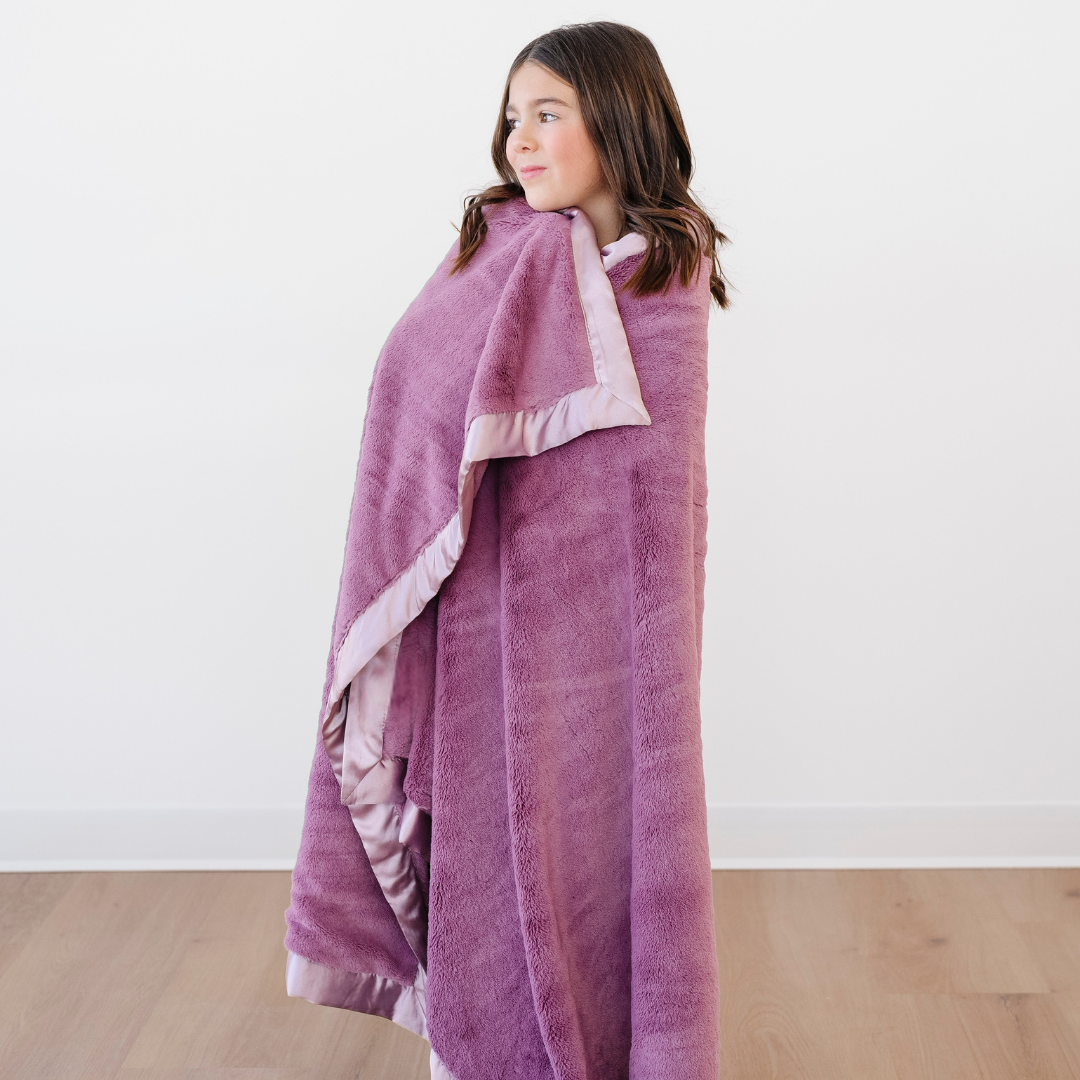 An adorable little girl is wrapped in a light purple blanket that is a toddler blanket and a lush blanket. This soft blanket has a cute satin border as well. This luxury blanket is a Saranoni blanket and is a great toddler gift or little girl gift. 