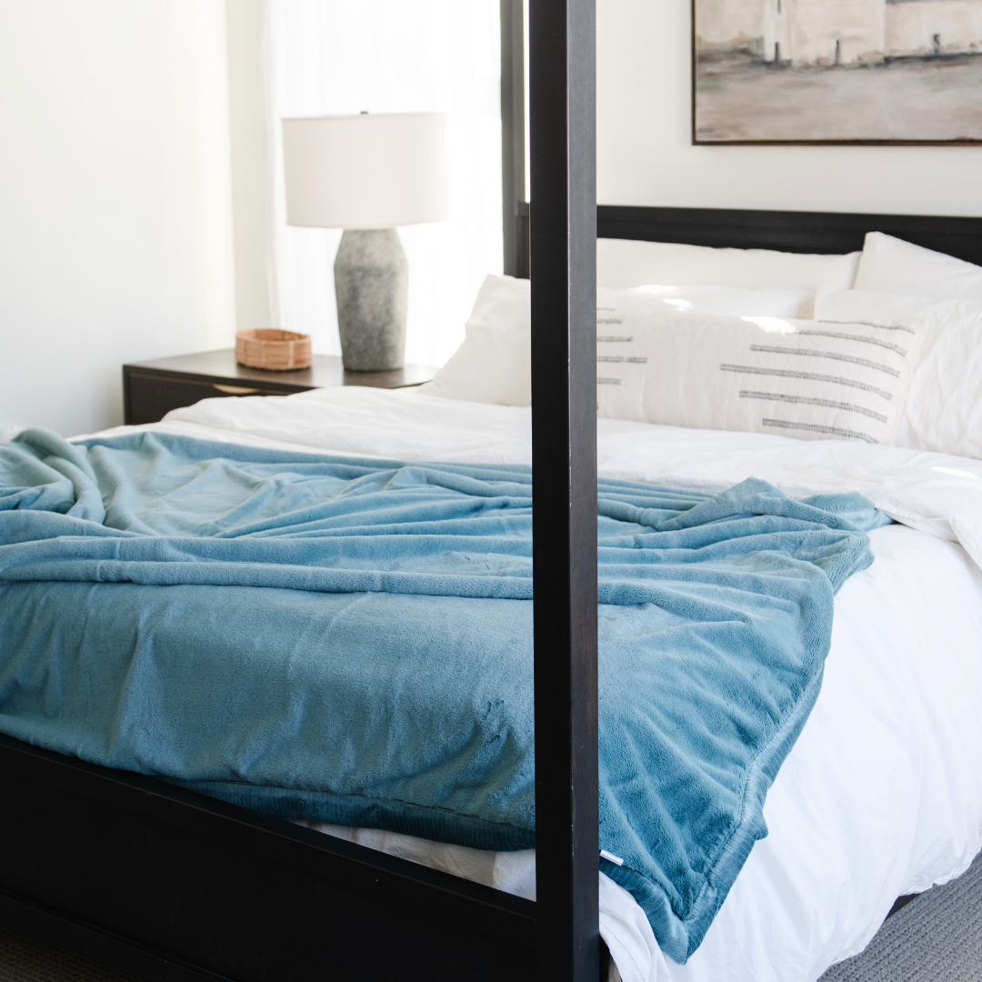 On a bed there is a Mineral Blue Lush XL Saranoni blanket, designed for oversized comfort and featuring a buttery soft, plush texture. This luxurious Saranoni blanket is perfect for cozy evenings, reading, or home decor. The Saranoni blanket adds warmth, style, and unmatched softness, making it an ideal gift or essential for ultimate relaxation. The soft blanket is a luxury blanket and a Saranoni blanket. 