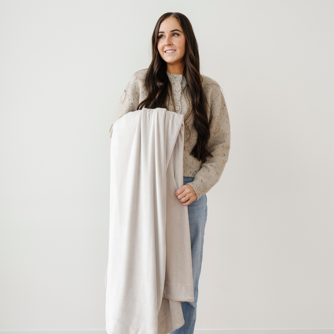 A smiling woman is holding a neutral minky stretch blanket that is an extra large blanket. This Saranoni blanket is very stretchy, durable, and breathable. This cozy blanket is great for gifting, lounging, or on the go. 