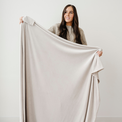 A woman is holding a neutral minky stretch blanket. This soft blanket is a Saranoni luxury blanket that is very stretchy, durable, and breathable. Enjoy this cozy blanket for years to come. 