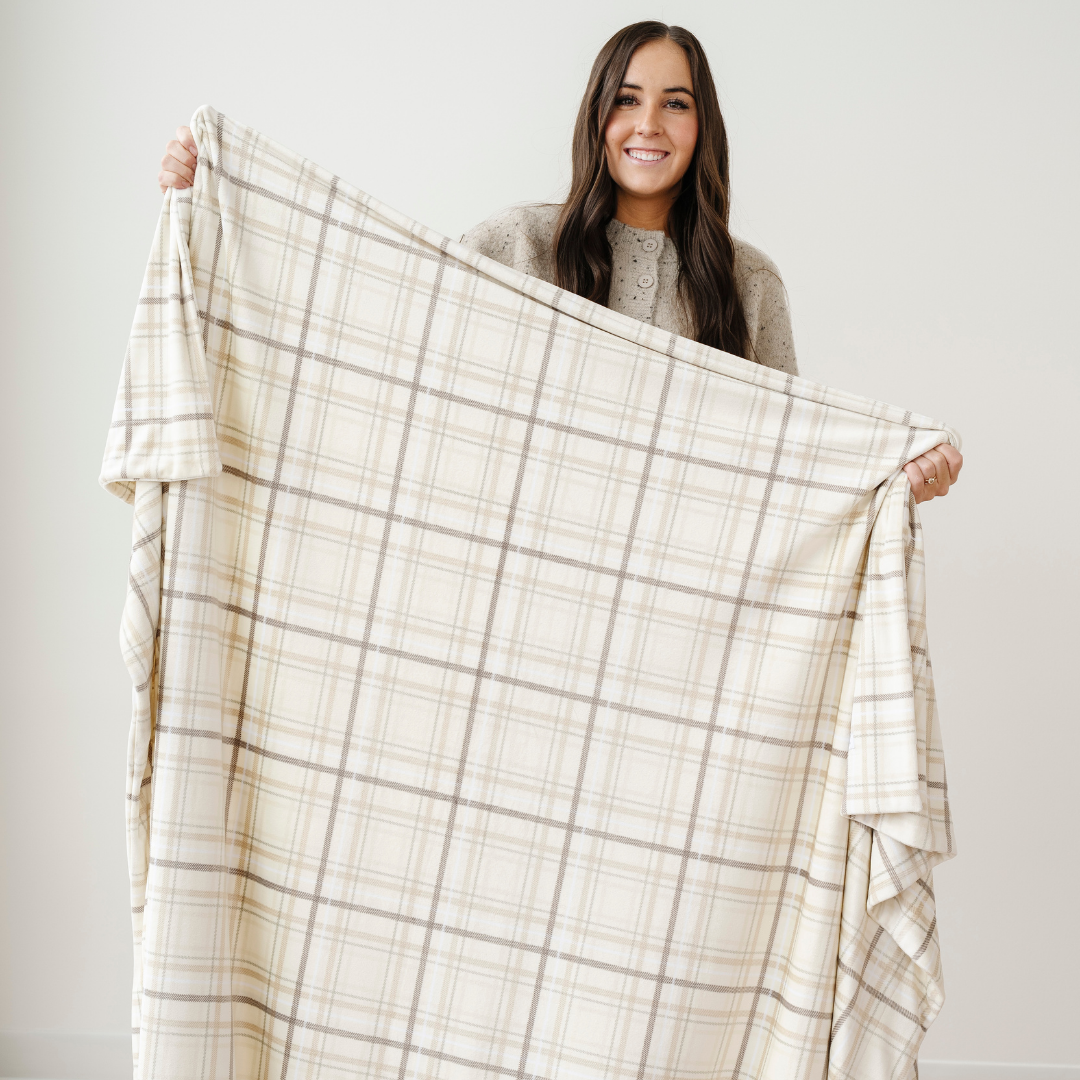 A smiling woman is holding up a neutral plaid minky stretch blanket. This soft blanket is a luxury blanket and a Saranoni blanket. This blanket is very stretchy, breathable, and durable. Enjoy this throw blanket for years to come!
