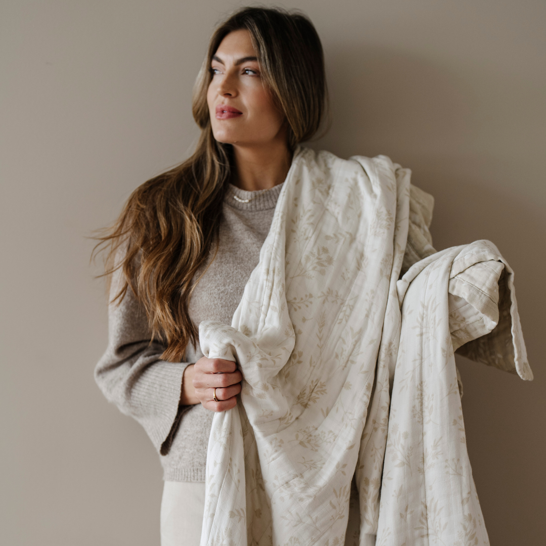 A woman holds a white and tan bamboo rayon muslin quilt that has a floral pattern on it. This soft blanket is a luxury blanket and a Saranoni blanket. This blanket is great for gifting, relaxing, and as a home decor staple. Enjoy this cozy blanket for years to come. 