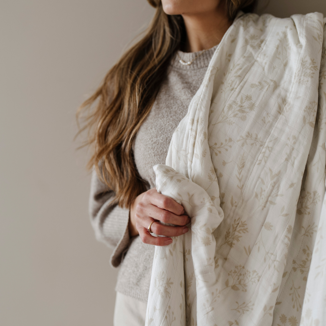 A woman is holding a bamboo rayon muslin quilt that is white and tan with a floral pattern. This soft blanket is great for gifting, relaxing, or as a home decor piece.  This Saranoni blanket is an extra-large blanket. 