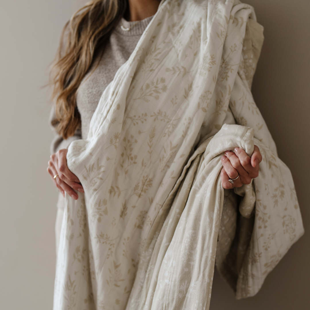 A woman is holding a white and tan floral patterned bamboo rayon muslin quilt. This Saranoni blanket is a soft blanket and a luxury blanket. This cozy blanket is a great comfort gift, relaxation item, or home decor staple. 