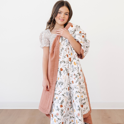 A smiling little girl holds a Saranoni blanket that is a quilt that has flowers on it. This soft blanket is a luxury blanket and is a muslin fabric. This quilt is a great kids blanket and an adorable baby gift, toddler gift, or addition to a nursery or bedroom. Enjoy this cozy quilt for years to come!