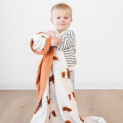 A little boy holds a bamboo rayon muslin quilt with a highland cow pattern on it. The soft quilt is a Saranoni blanket and is an adorable addition to a bedroom or playroom. This cozy blanket also makes a great baby gift or toddler gift. 