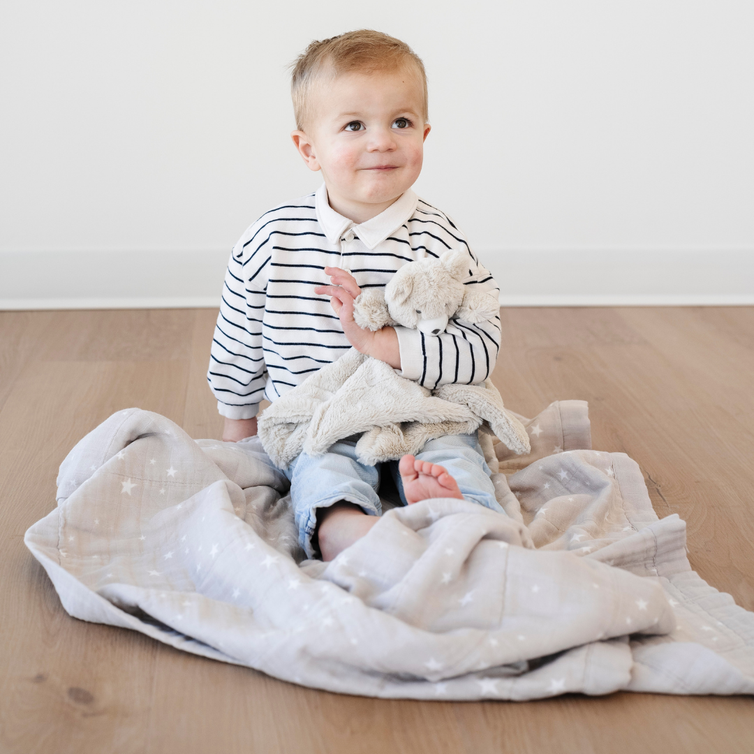 A little boy is sitting on a bamboo rayon muslin quilt that has a little star pattern on it and is holding a bear stuffed animal lovey. This soft quilt is a great kid's blanket and a Saranoni blanket. The muslin fabric is softer with every wash, making it a great baby gift or toddler gift. Enjoy this soft blanket for years to come. 