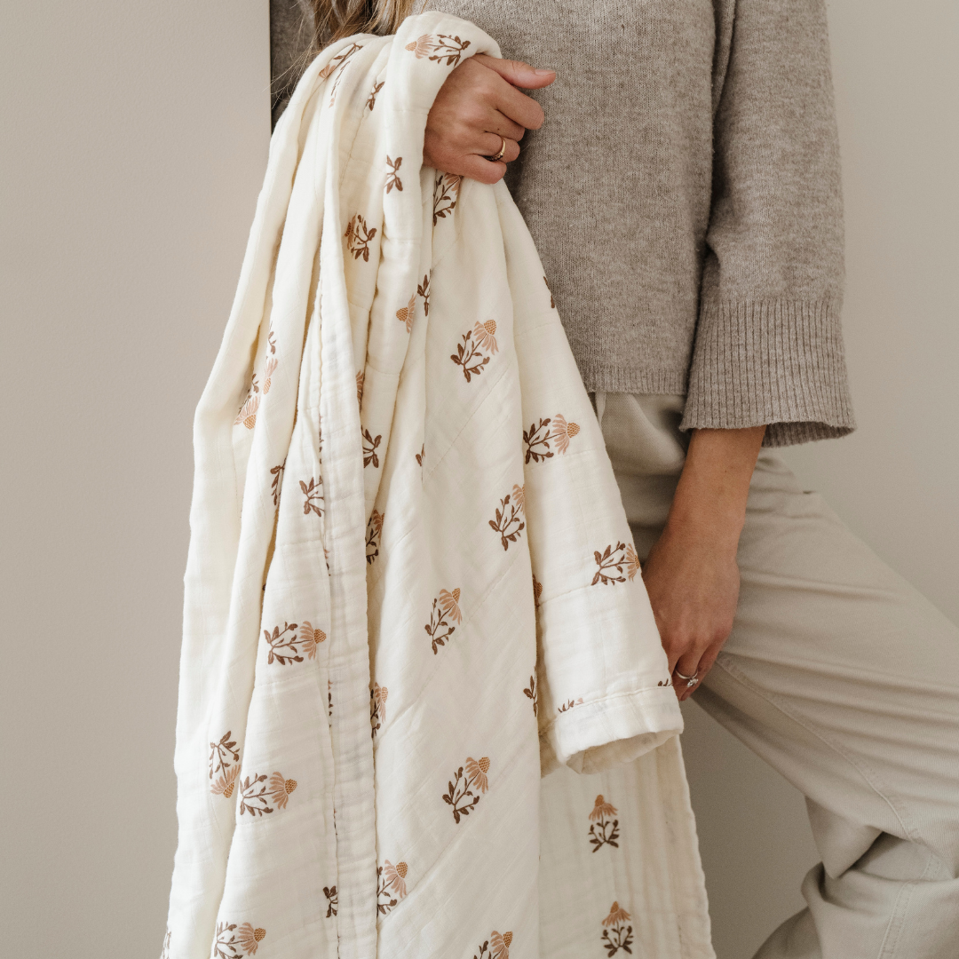 A woman holds a floral patterned bamboo rayon muslin quilt. This soft blanket get softer with every wash and is a Saranoni blanket. This blanket is a great comfort gift, relaxing blanket, or home decor staple. 