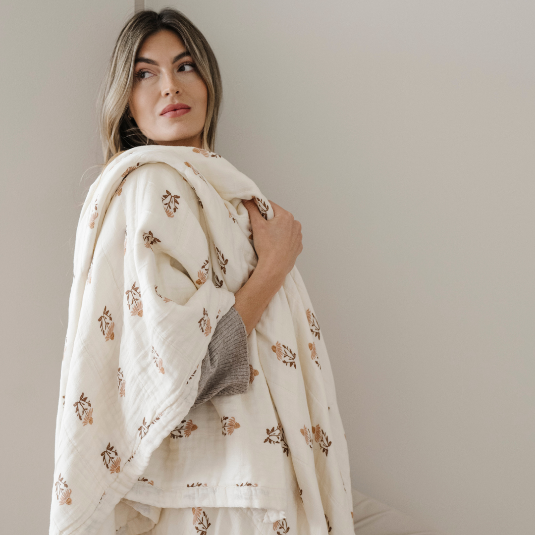 A woman looks off into the distance. She is holding a bamboo rayon muslin quilt. This soft blanket is a luxury blanket and a Saranoni blanket with a beautiful floral pattern. Elevate your home decor with this cozy blanket!