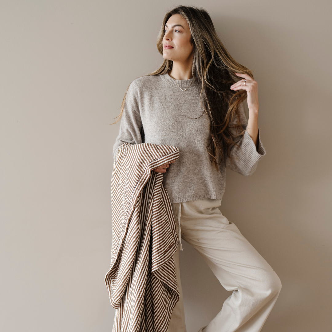 A woman is holding a bamboo rayon muslin quilt that has a brown and white striped pattern on it. This soft blanket is a luxury blanket and a Saranoni blanket. This muslin quilt gets softer with every was and is great for relaxing, gifting, and styling in your home. 