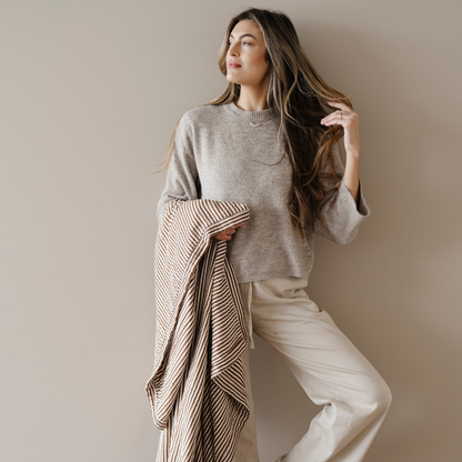 A woman is holding a bamboo rayon muslin quilt that has a brown and white striped pattern on it. This soft blanket is a luxury blanket and a Saranoni blanket. This muslin quilt gets softer with every was and is great for relaxing, gifting, and styling in your home. 