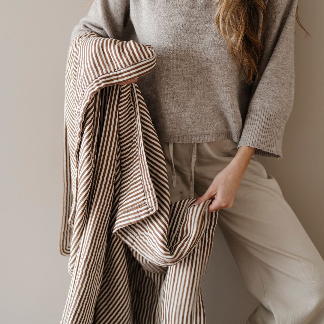 A woman is holding a brown and white striped bamboo rayon muslin quilt that is a Saranoni blanket. This luxury blanket gets softer with every was and is great for home decor, relaxing, and gifting. Enjoy this cozy blanket for years to come!
