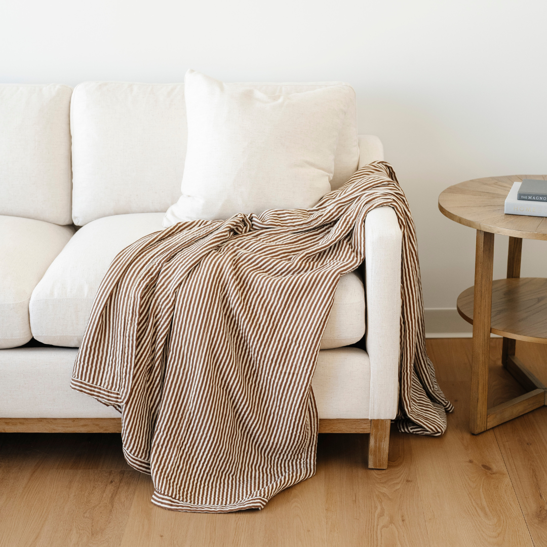 A brown and white striped bamboo rayon muslin blanket is draped on a couch. This soft blanket is a luxury blanket and a Saranoni blanket. Use this blanket to gift to someone, relax in, or as a home decor staple. 