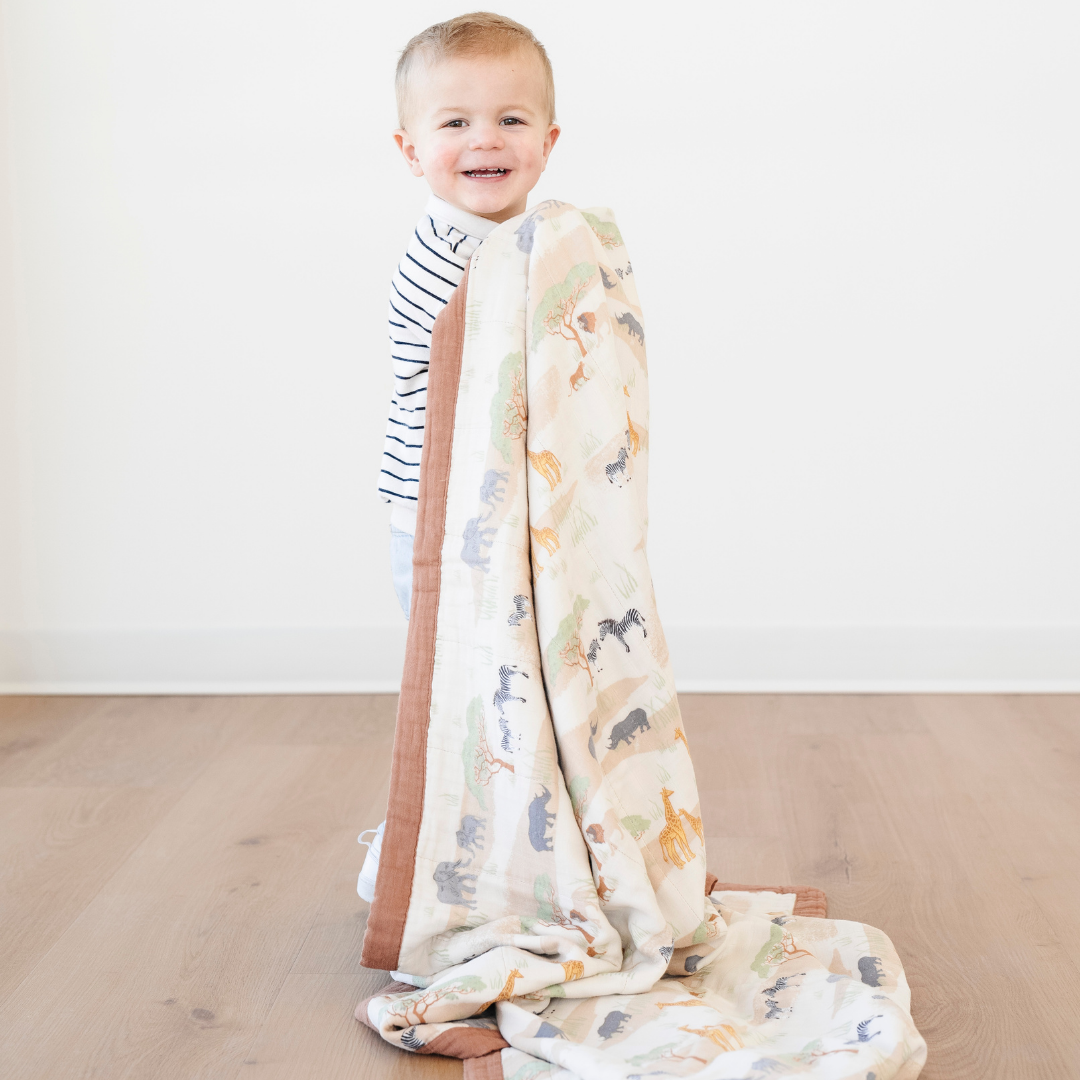 A smiling little boy is holding bamboo rayon muslin quilt that has a safari animal pattern on it. This soft blanket is a luxury blanket and a Saranoni blanket. The soft muslin fabric makes it a great baby gift, gift for toddlers, or addition to a bedroom or nursery. 