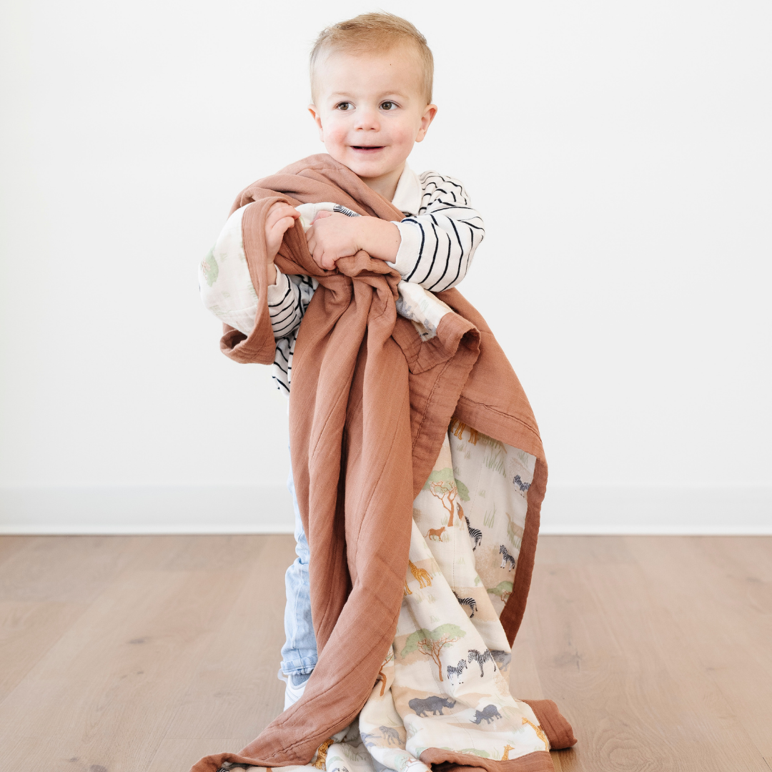 A little boy is hugging a bamboo rayon muslin quilt with a safari animal pattern on it. This soft blanket is a Saranoni blanket. This a great toddler gift or baby gift. 
