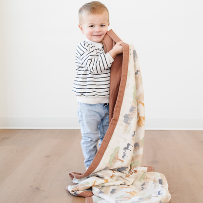 A little boy is holding a bamboo rayon muslin quilt with safari animals on it. This soft blanket is a Saranoni blanket and is a great kids blanket. This is a great baby gift or toddler gift because of the soft muslin fabric. 