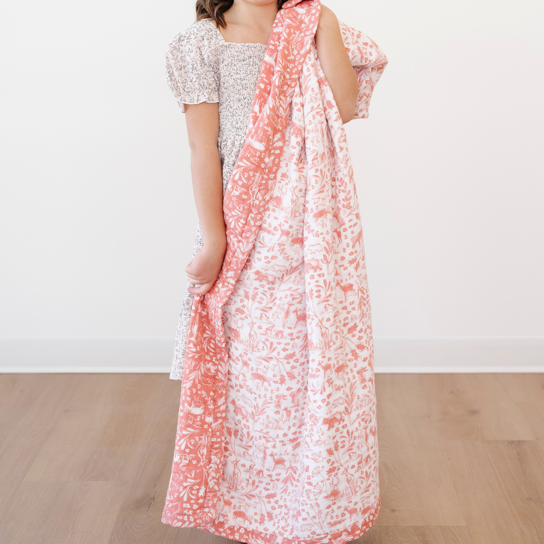 A little girl holds a pink and white bamboo rayon muslin quilt that has a woodland animal pattern on it. This soft blanket is a Saranoni blanket and is a great kids blanket. Since the fabric gets softer with every wash it is a great baby gift or toddler gift. 