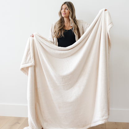 A woman holds a Natural (cream) Lush XL Saranoni blanket, designed for oversized comfort and featuring a buttery soft, plush texture. This luxurious Saranoni blanket is perfect for cozy evenings, reading, or home decor. The Saranoni blanket adds warmth, style, and unmatched softness, making it an ideal gift or essential for ultimate relaxation.