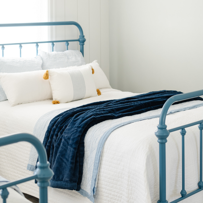 On a bed with white and light blue colors is a Navy Lush XL Saranoni blanket, designed for oversized comfort and featuring a buttery soft, plush texture. This luxurious Saranoni blanket is perfect for cozy evenings, reading, or home decor. The Saranoni blanket adds warmth, style, and unmatched softness, making it an ideal gift or essential for ultimate relaxation.