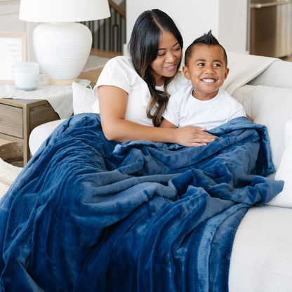 A mother and her son sit under a Navy Lush XL Saranoni blanket, designed for oversized comfort and featuring a buttery soft, plush texture. This luxurious Saranoni blanket is perfect for cozy evenings, reading, or home decor. The Saranoni blanket adds warmth, style, and unmatched softness, making it an ideal gift or essential for ultimate relaxation.
