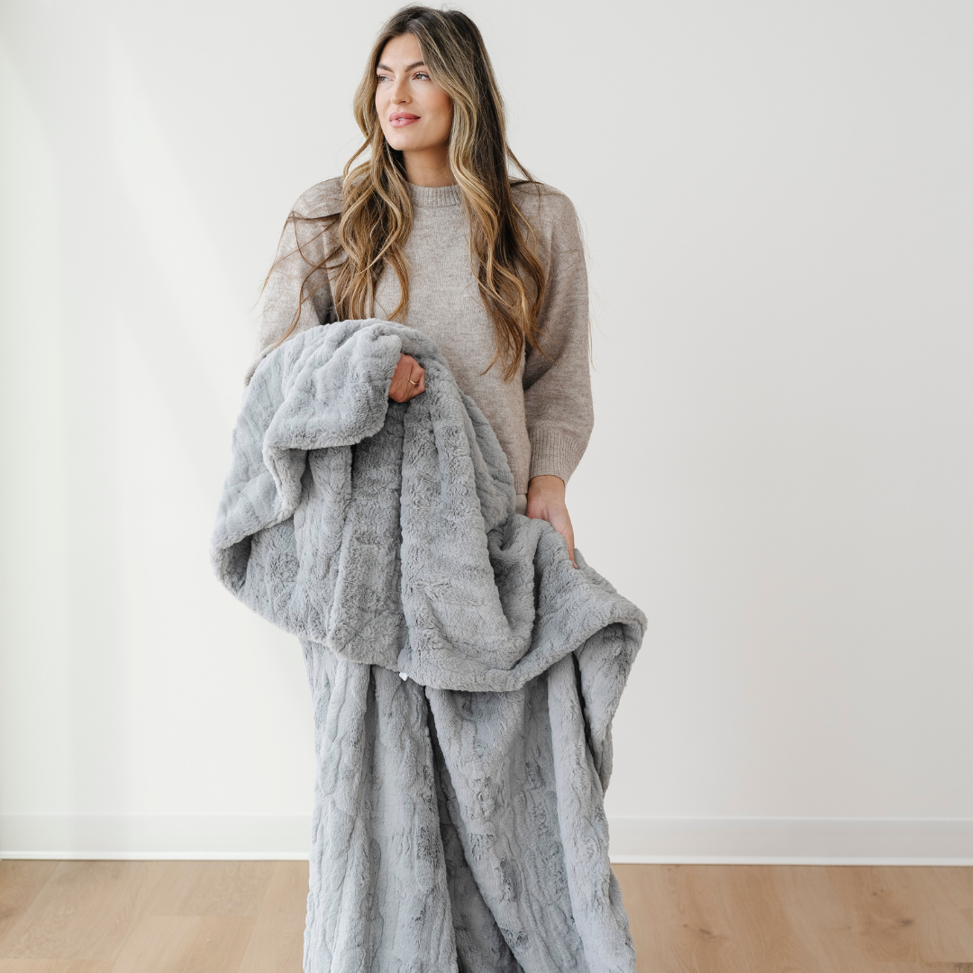 A woman holds a gray patterned faux fur blanket that is a throw blanket. This blanket is also a Saranoni blanket that is very durable and soft. Use this blanket for gifting, relaxing, or home decor. 