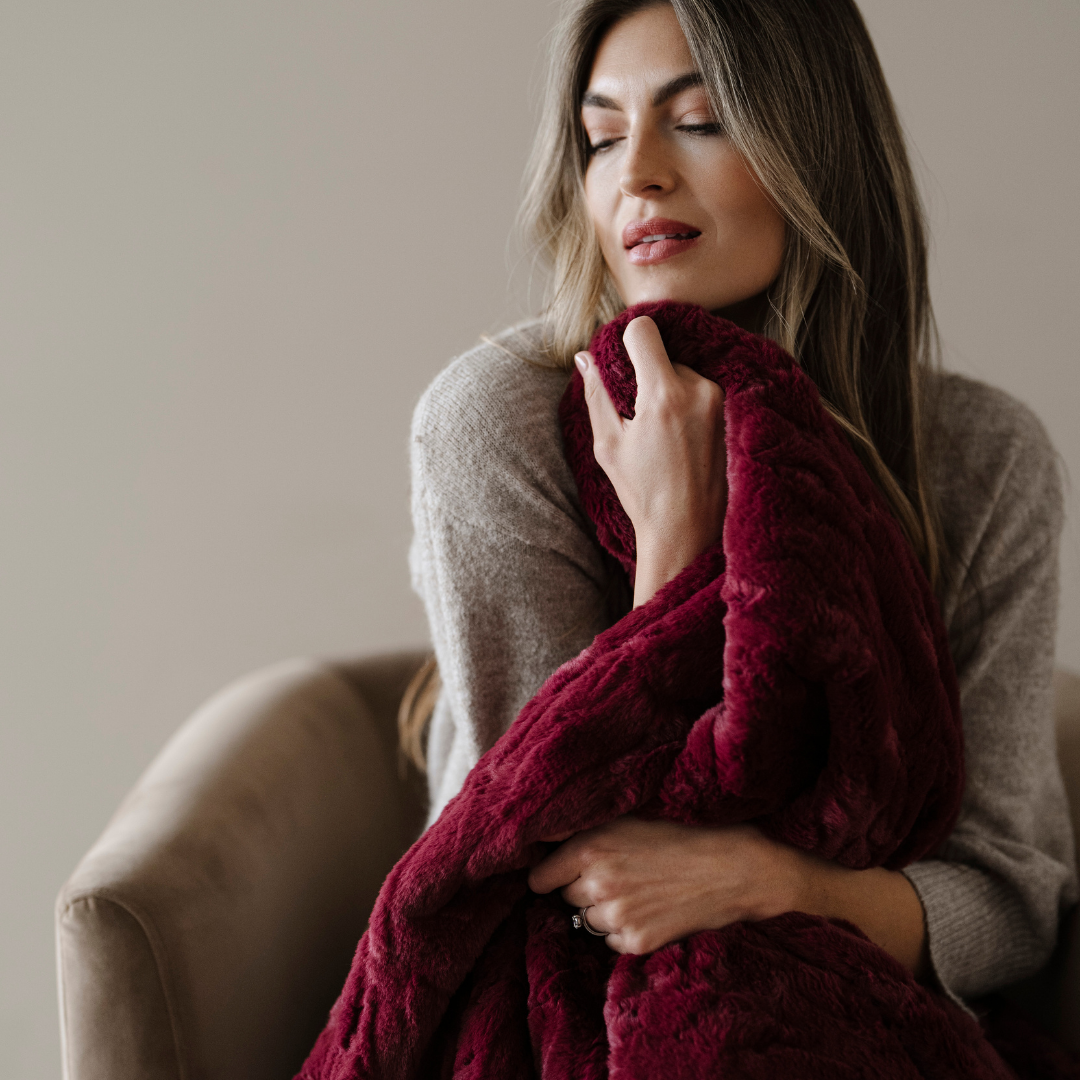 A woman is holding a deep red patterned faux fur blanket. This soft blanket is a Saranoni blanket and is great for luxury living. 