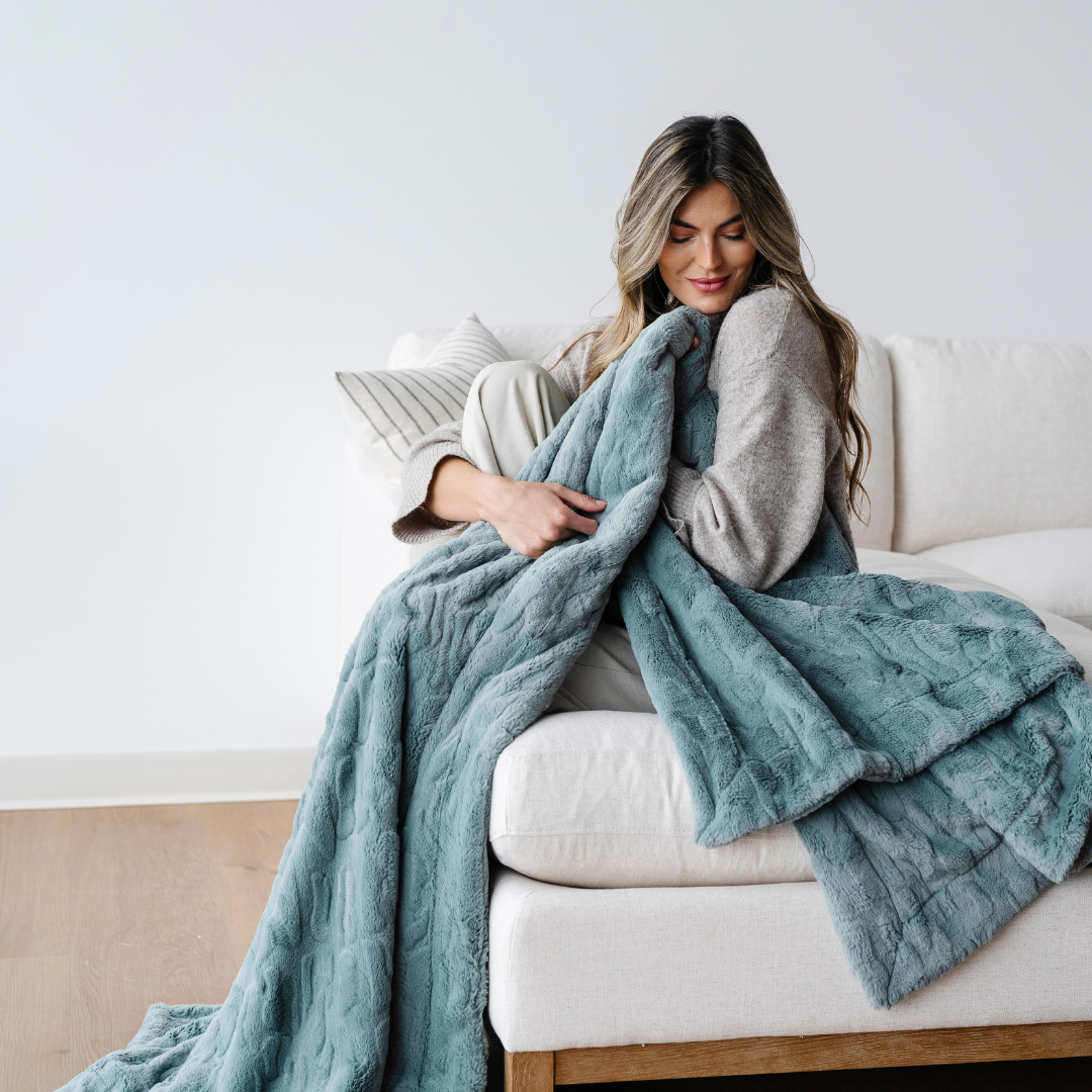 A woman is sitting on a couch and holding a eucalyptus patterned faux fur blanket. This soft blanket is perfect for luxury living, gifting, and home decor. This Saranoni blanket has a luxurious texture and is an extra large throw blanket. 
