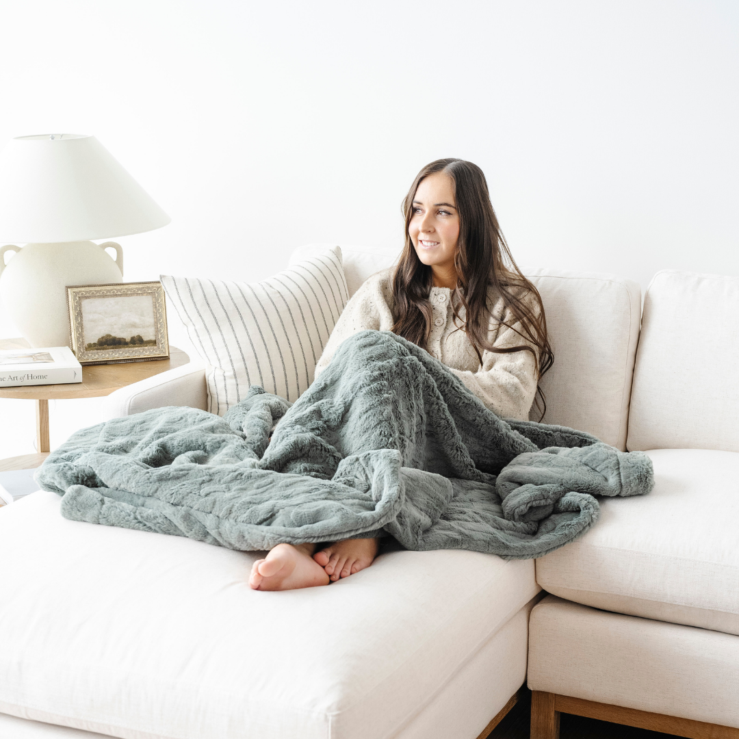 A woman sits on a white couch with a green patterned faux fur blanket. This soft blanket is a great home decor staple and is also a cozy comfort gift and great for relaxing. This Saranoni blanket is durable and will last for years to come!