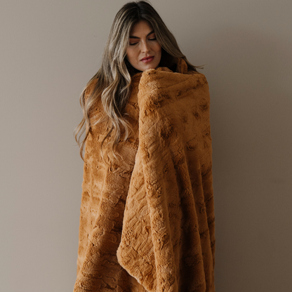 A woman is wrapped in a Saranoni blanket that is a copper colored patterned faux fur blanket. This throw blanket is perfect for home decor, adding to a luxury living room, or refreshing a bedroom. Enjoy this soft blanket for years to come. 