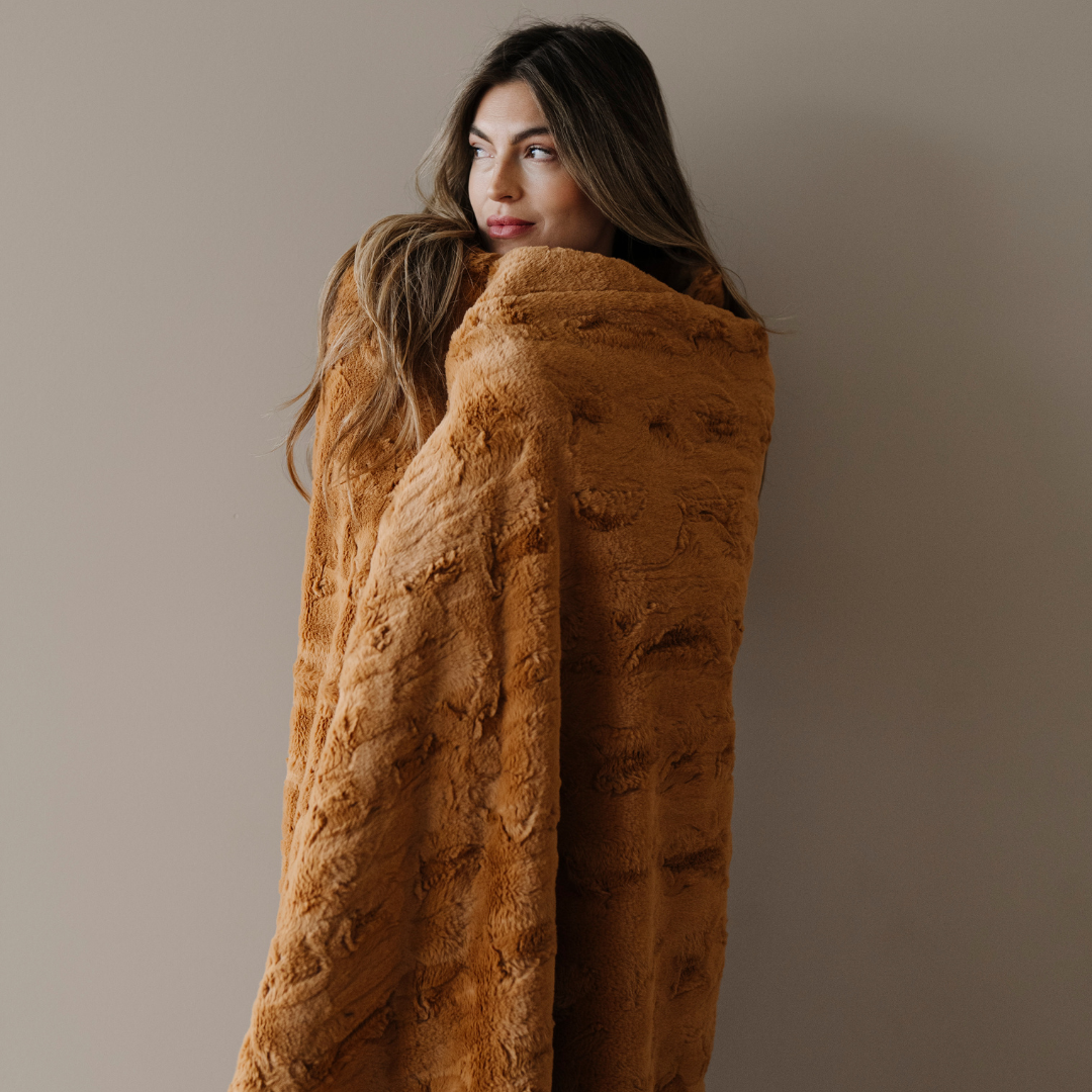 A woman is wrapped in a copper colored patterned faux fur blanket. This luxury blanket is a Saranoni blanket that is durable, soft, and luxurious. This blanket is perfect for gifting, lounging, or as home decor. 