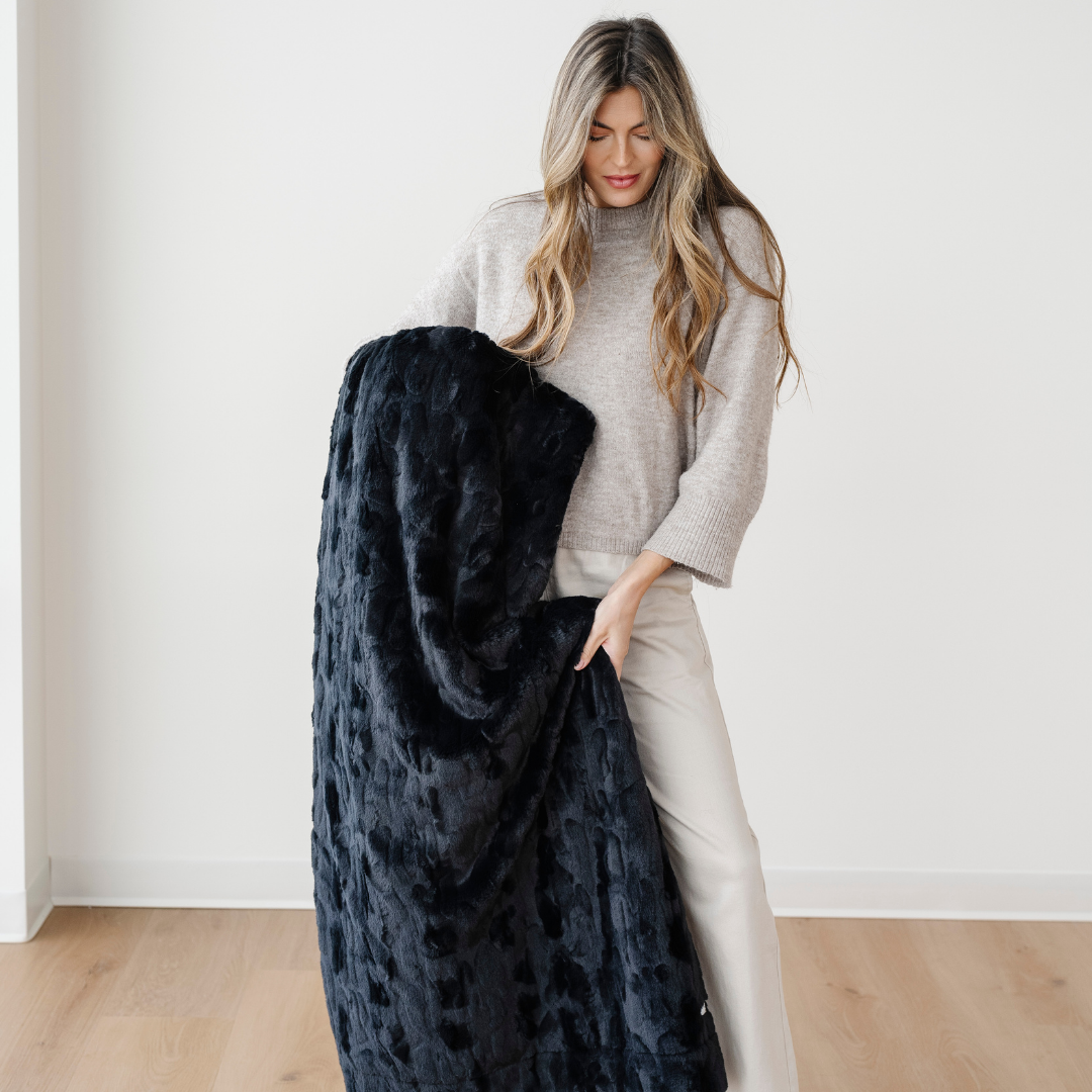 A woman is holding a black patterned faux fur blanket. This soft blanket is a luxury blanket. This blanket is perfect for gifting, lounging, and home decor. Enjoy luxury living with this blanket for years to come. 