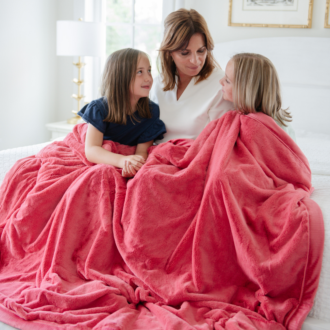 A mother and her two daughters sit on a bed under a Primrose (deep red/pink) Lush XL Saranoni blanket, designed for oversized comfort and featuring a buttery soft, plush texture. This luxurious Saranoni blanket is perfect for cozy evenings, reading, or home decor. The Saranoni blanket adds warmth, style, and unmatched softness, making it an ideal gift or essential for ultimate relaxation.