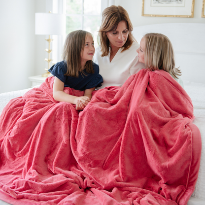 A mother and her two daughters sit on a bed under a Primrose (deep red/pink) Lush XL Saranoni blanket, designed for oversized comfort and featuring a buttery soft, plush texture. This luxurious Saranoni blanket is perfect for cozy evenings, reading, or home decor. The Saranoni blanket adds warmth, style, and unmatched softness, making it an ideal gift or essential for ultimate relaxation.