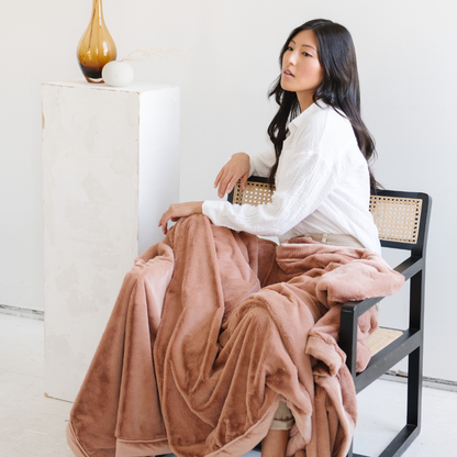 A woman sits in a chair and sits under a Rosy (dusty peach) Lush XL Saranoni blanket, designed for oversized comfort and featuring a buttery soft, plush texture. This luxurious Saranoni blanket is perfect for cozy evenings, reading, or home decor. The Saranoni blanket adds warmth, style, and unmatched softness, making it an ideal gift or essential for ultimate relaxation.