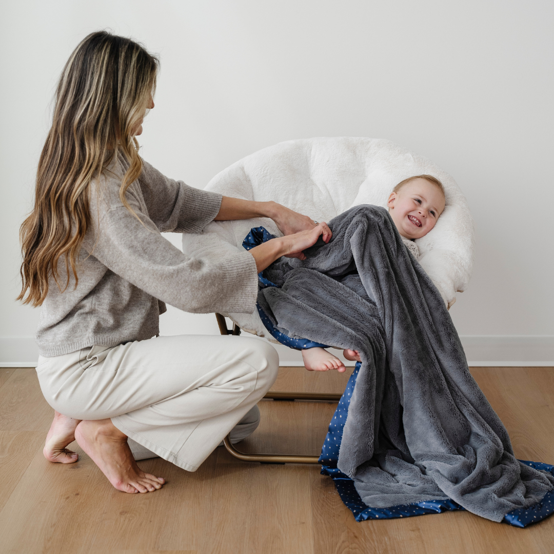 A mother is by her little boy who is sitting in a saucer chair. He has a gray lush blanket that has a star satin border. This soft blanket is a luxury blanket and a Saranoni blanket. This toddler blanket is also a great toddler gift and addition to a kid's bedroom. 