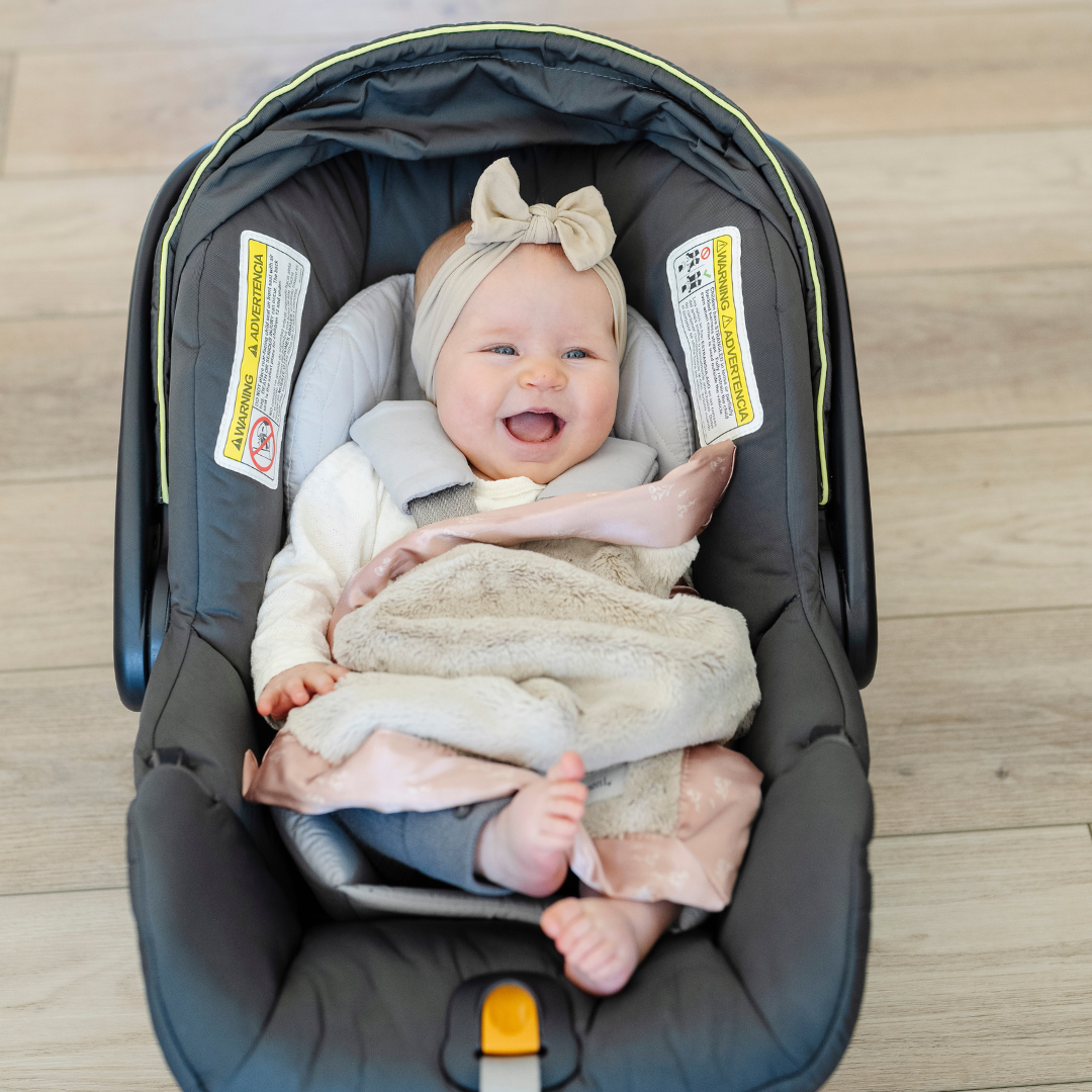 A baby girl in her carseat with a satin border blanket. The lush blanket is tan with a peach satin border that has a floral design on it. The satin border blanket is a lush blanket with a satin border. The soft blanket is a luxury blanket and a Saranoni blanket. This mini blanket is an adorable baby blanket and perfect for littles. 