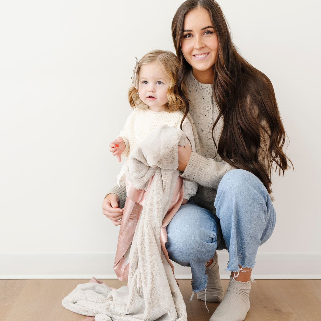 A mother is by her little girl. They are holding a light tan lush blanket that has a floral satin back and border. The soft blanket is a luxury blanket and a Saranoni blanket. Enjoy this cozy blanket as a toddler gift or a little girl gift. 