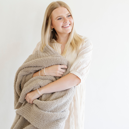 A woman smiles and holds a Bamboni® blanket by Saranoni. This ultra-soft blanket is a taupe blanket and made from a premium blend of bamboo rayon and poly microfiber, offering a lightweight, breathable, and fluffy feel. Perfect as a throw blanket, luxury blanket, plush throw, or soft blanket, it adds warmth and elegance to any living room, bedroom, or home décor setting. Designed for ultimate comfort, this buttery-soft Saranoni blanket is ideal for gifting. Enjoy this cozy blanket for years to come!