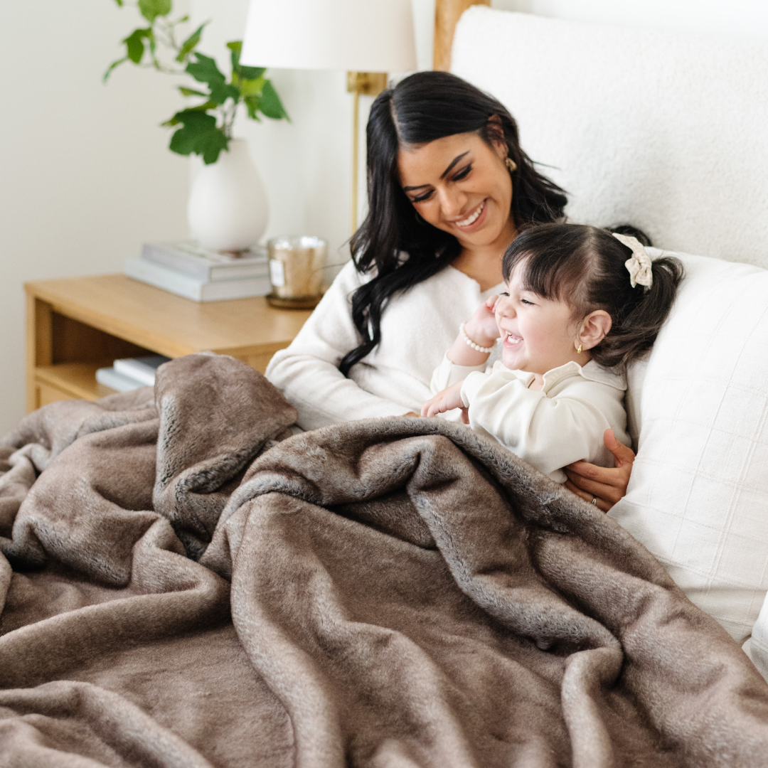 A mother and her daughter sit under a Saranoni Grand Faux Fur blanket, featuring an elegant light brown tone and luxuriously soft texture. This cozy, queen-sized blanket and faux fur blanket offers soothing weight, premium softness, timeless comfort, and is a soft blanket. Perfect as a big blanket and luxury blanket for snuggling or as a luxury bedroom accent, this Saranoni blanket elevates every cozy moment and is a cozy blanket.