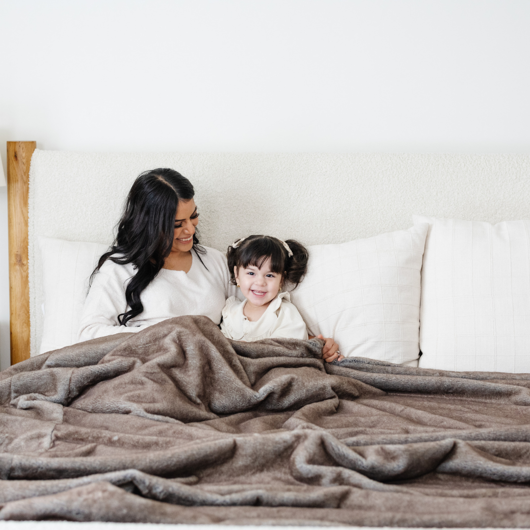 A mother and her daughter smile and sit under a Saranoni Grand Faux Fur blanket, featuring an elegant light brown tone and luxuriously soft texture. This cozy, queen-sized blanket and faux fur blanket offers soothing weight, premium softness, timeless comfort, and is a soft blanket. Perfect as a big blanket and luxury blanket for snuggling or as a luxury bedroom accent, this Saranoni blanket elevates every cozy moment and is a cozy blanket.